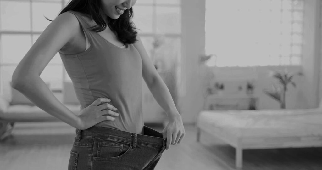 A woman smiling and holding out the waistband of her jeans.