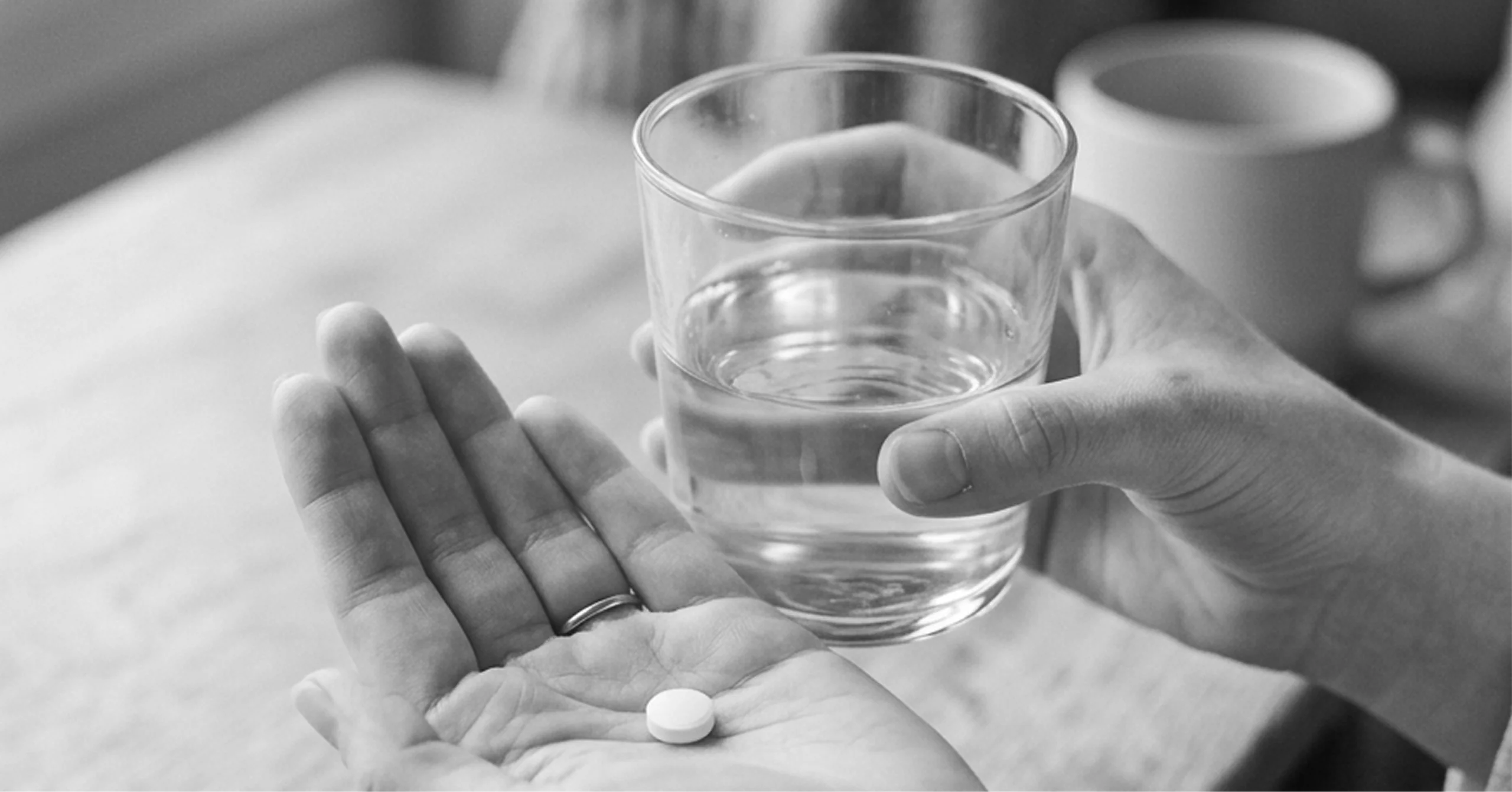 A person holding a single round tablet in one open palm and a glass of water in the other hand, above a table.