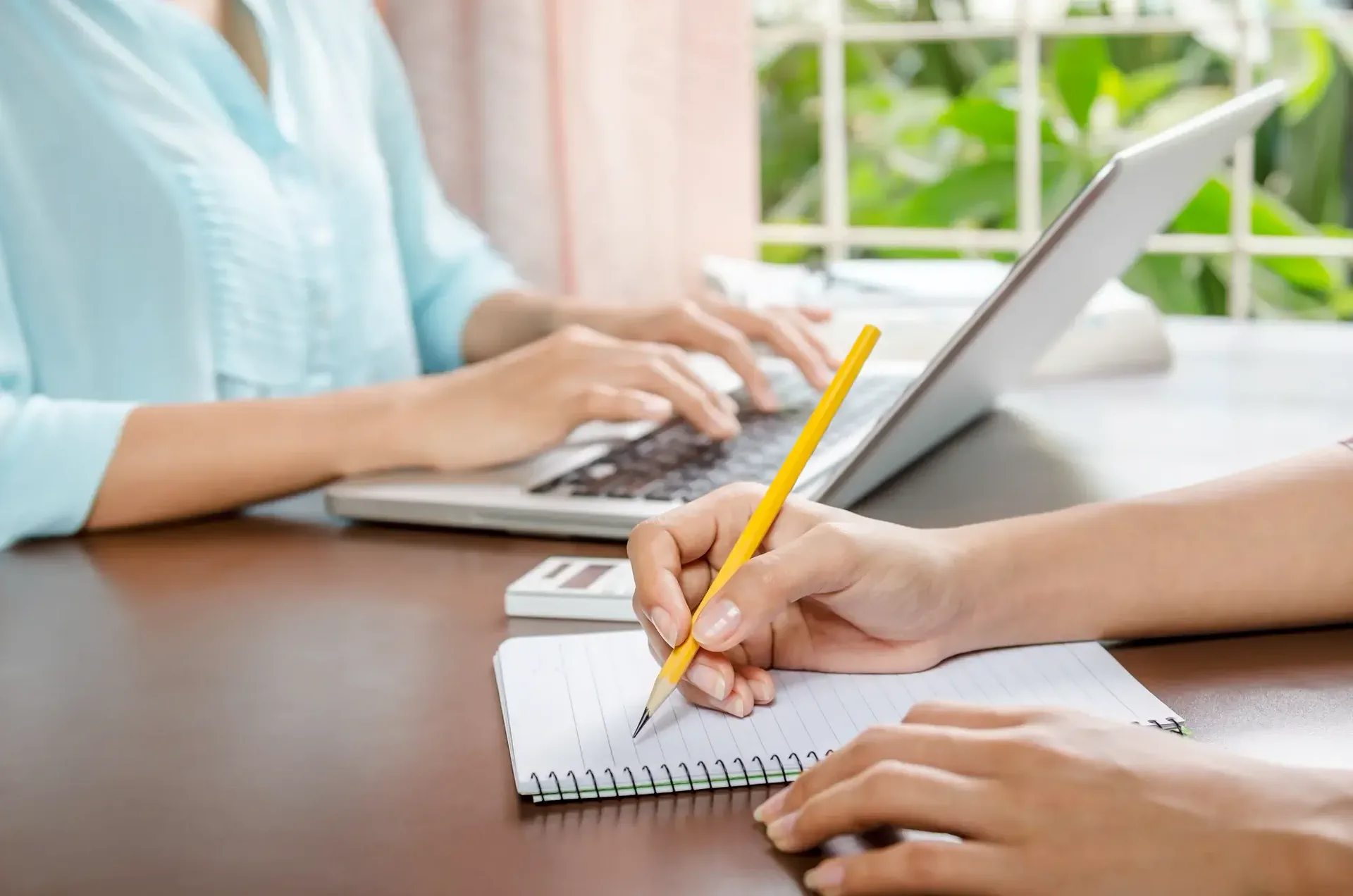 Two people work on a desk, one typing on a laptop and the other writing in a notebook.