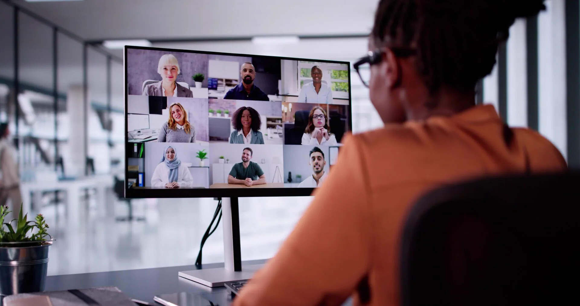 Person attending an online webinar, with nine participants shown on a desktop monitor in an office